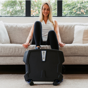 Female is setting on her couch while exercising on the Helix Thera Scoop Lateral Elliptical Pedaler